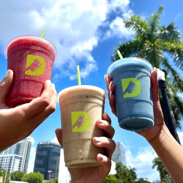Three hands hold Diced smoothies (red, tan, blue) against a sunny sky with palm trees and buildings.