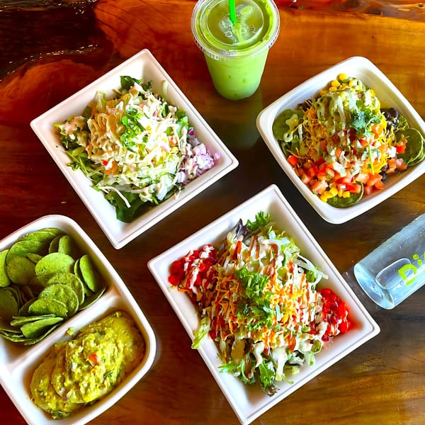 Four healthy Diced salads, green smoothie, chips, guacamole, and a water bottle on a wooden table.