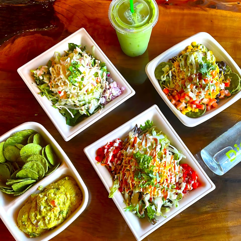 Four healthy Diced salads, green smoothie, chips, guacamole, and a water bottle on a wooden table.