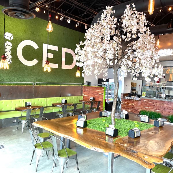 Diced restaurant interior with green turf wall, large wooden tables, and a decorative white blossom tree.