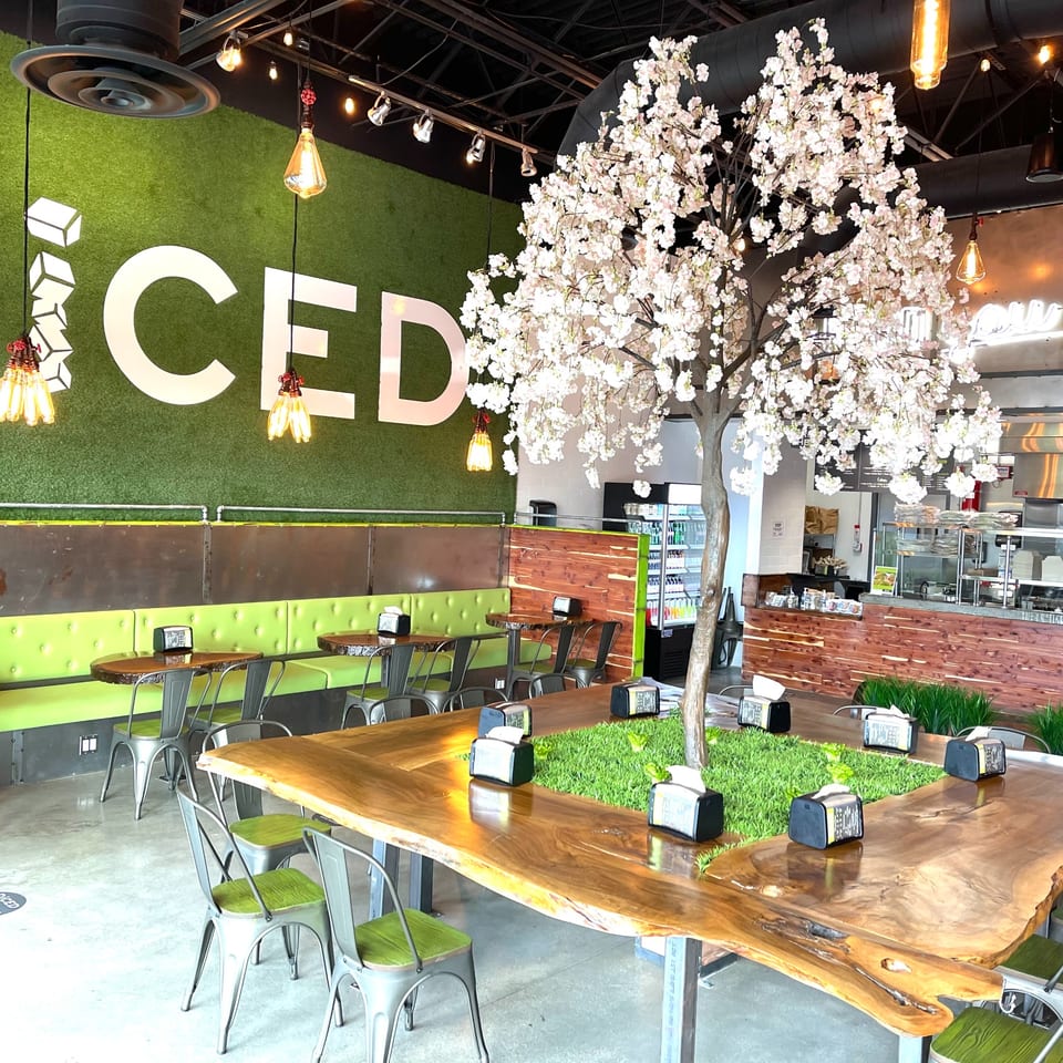 Diced restaurant interior with green turf wall, large wooden tables, and a decorative white blossom tree.