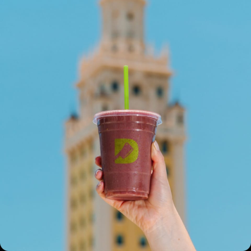 A hand holds a Diced smoothie with a green straw against a blue sky and blurred building.