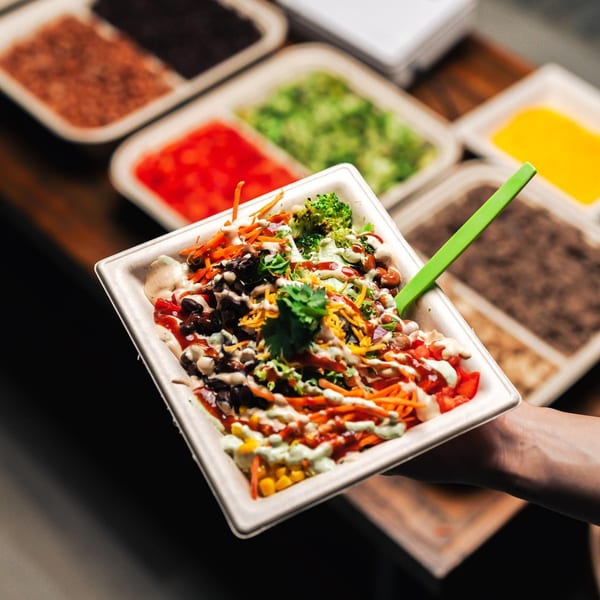 A Diced healthy bowl with broccoli, carrots, beans, corn, and sauce, held above ingredient trays.