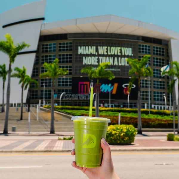 Hand holding a Diced green smoothie with the FTX Arena and palm trees in the background.