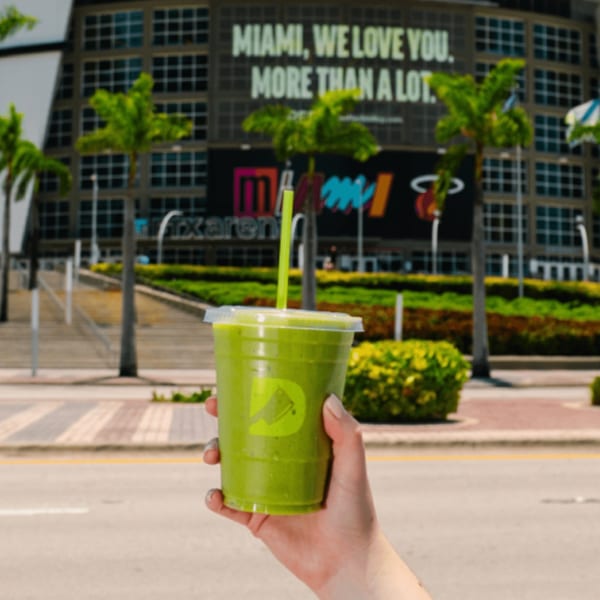 Hand holding a Diced green smoothie with a Miami arena and palm trees in the background.
