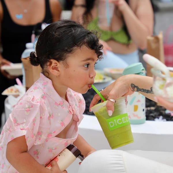 A young child in a pink dress sips a green smoothie from Diced through a straw.