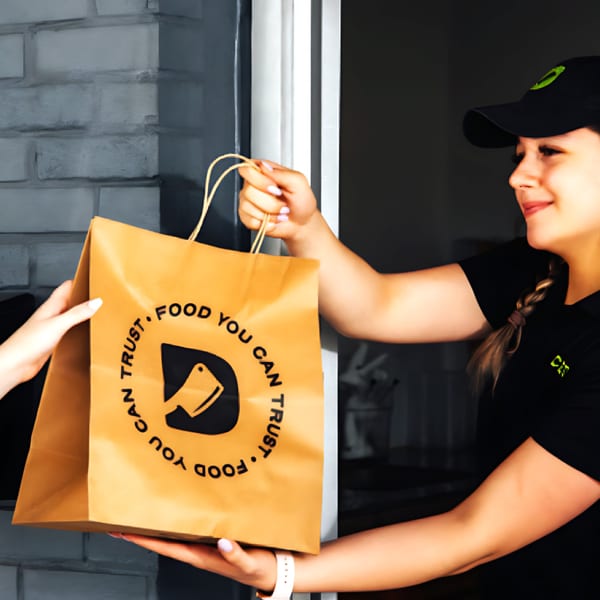 A Diced employee in a black cap hands a Diced branded brown paper bag to a customer.