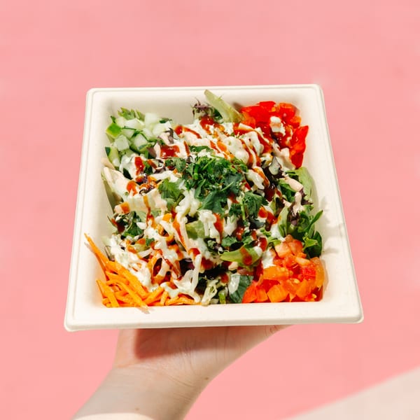 A hand holds a Diced healthy salad bowl with fresh vegetables and sauces against a pink background.