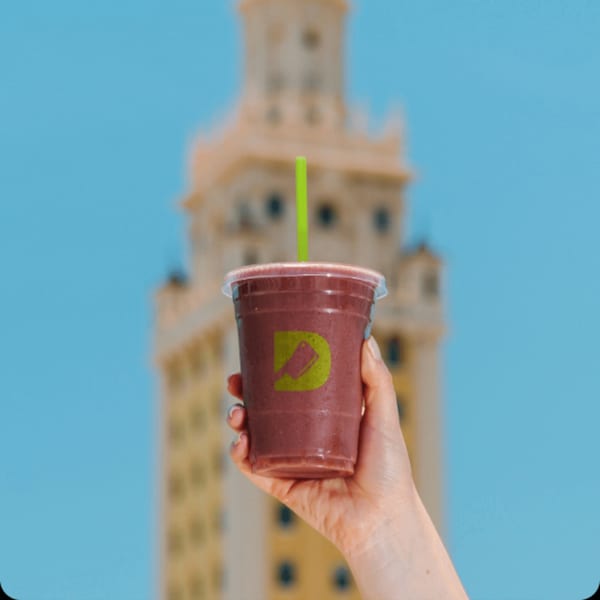 Hand holding a Diced berry smoothie with a green straw against a blurred building and blue sky.
